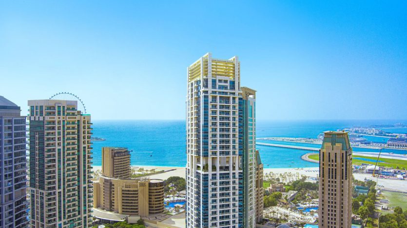 Vue aérienne d&#039;une ville côtière avec des immeubles modernes de grande hauteur avec l&#039;océan et une grande roue visible en arrière-plan sous un ciel bleu clair, parfaite pour mettre en valeur l&#039;immobilier Dubaï.