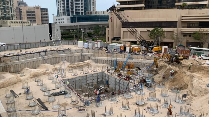 Un chantier de construction animé avec des ouvriers à Dubaï, entouré d&#039;immeubles de grande hauteur. Excavation et piliers structurels en cours, avec une grue et divers véhicules de construction visibles.