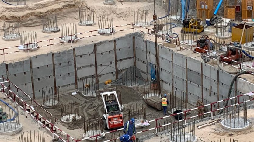 Un chantier de construction avec des ouvriers à Dubaï, comprenant une grande fosse creusée renforcée par des murs en béton et des barres métalliques. Des engins et outils de construction sont visibles autour du site.