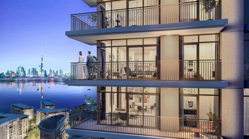 Un rendu architectural d&#039;un immeuble résidentiel moderne à plusieurs étages à l&#039;aube. Deux personnes sont visibles sur un balcon surplombant un paysage urbain avec un gratte-ciel important à Dubaï en arrière-plan.