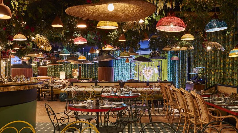 Un intérieur de restaurant vibrant et éclectique comprenant une gamme de lampes suspendues colorées, des chaises en osier et des tables dressées pour les repas, avec une verdure luxuriante et des motifs floraux sur les murs. Parfait pour les discussions sur l&#039;investissement