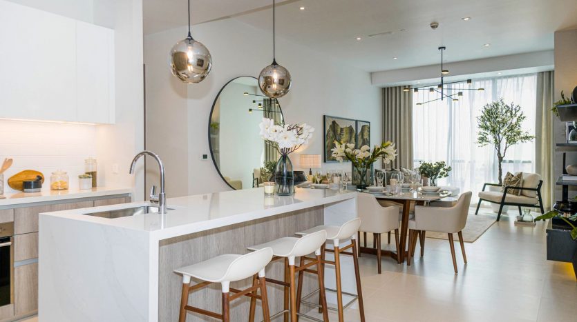 Cuisine et salle à manger modernes dans une villa de Dubaï avec un îlot blanc, des tabourets de bar, un grand miroir, des lampes argentées suspendues et une table à manger pour six personnes. Décor clair et aéré avec des éléments de design contemporain