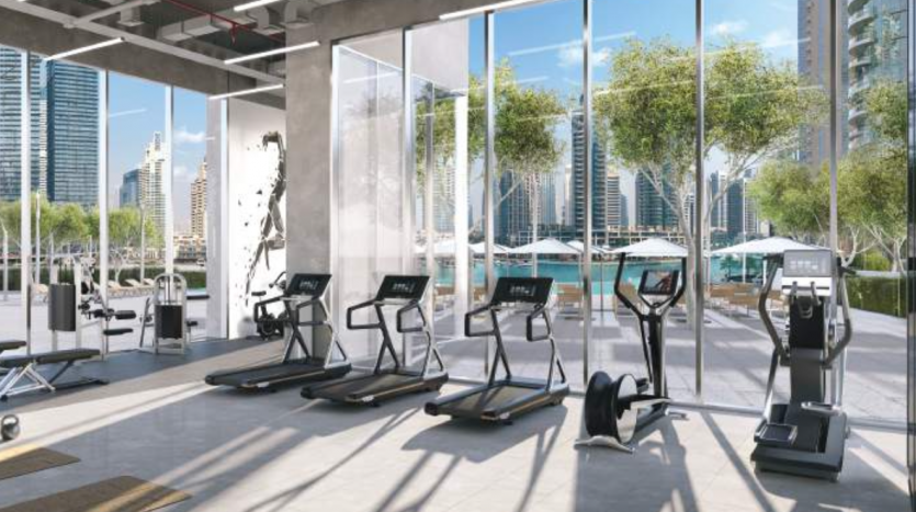Une salle de sport moderne avec des tapis roulants et des vélos elliptiques face à de grandes fenêtres donnant sur un paysage urbain ensoleillé avec des gratte-ciel et des palmiers, idéale pour ceux qui cherchent à investir à Dubaï.