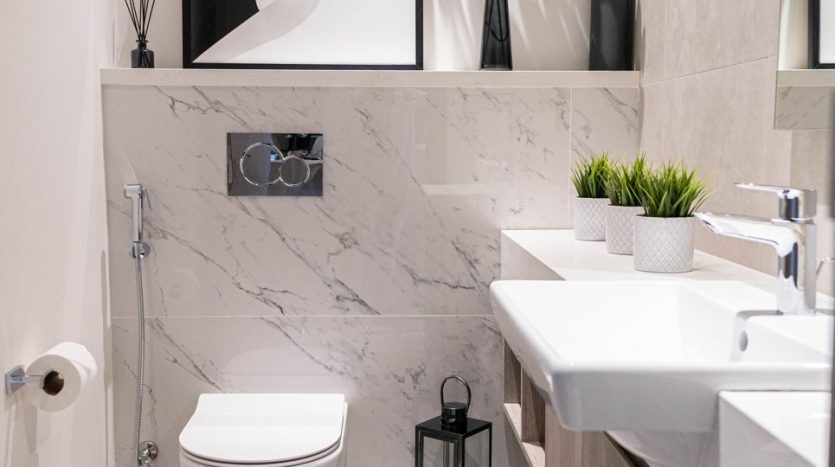 Une salle de bains moderne dans un appartement de Dubaï avec des murs en marbre élégants, comprenant des toilettes flottantes et un lavabo. Les éléments décoratifs comprennent une œuvre d&#039;art abstraite encadrée, des plantes vertes et des accessoires minimalistes.