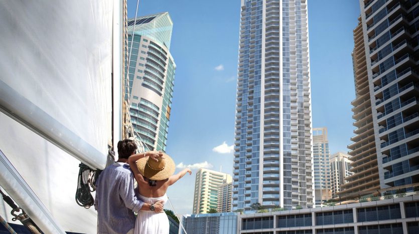 Un couple s&#039;embrasse sur un yacht, surplombant les toits d&#039;une ville moderne avec les imposants gratte-ciel de Dubaï et un ciel bleu clair. La femme tient un chapeau de soleil, indiquant une journée ensoleillée.