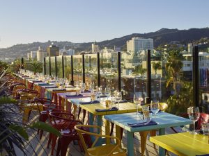 Terrasse extérieure du restaurant dotée de tables et de chaises colorées, surplombant un paysage urbain panoramique de Dubaï avec des collines en arrière-plan sous un ciel clair.