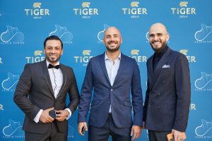 Trois hommes souriants en tenue d'affaires posent devant un fond bleu avec le logo Tiger Group lors d'un événement immobilier en plein air à Dubaï.