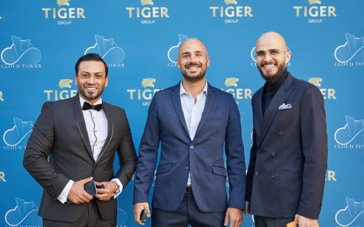 Trois hommes souriants en tenue d'affaires posent devant un fond bleu avec le logo Tiger Group lors d'un événement immobilier en plein air à Dubaï.