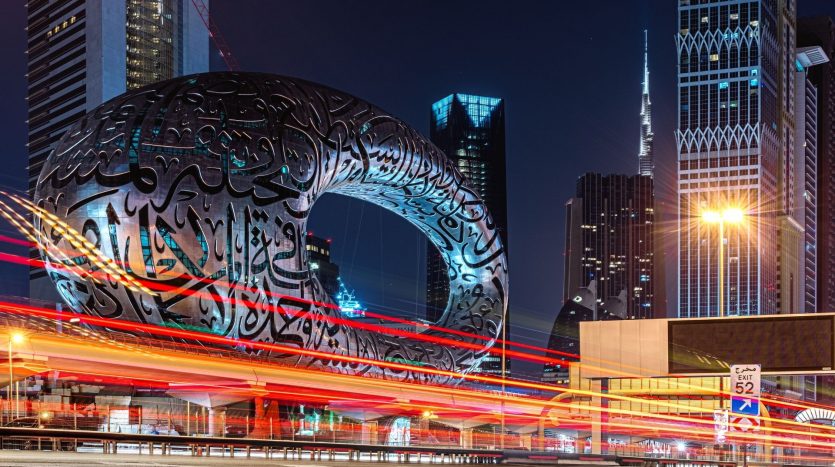 Paysage urbain nocturne avec le Musée du Futur de Dubaï, un bâtiment circulaire avec des motifs de calligraphie arabe complexes, sur fond de gratte-ciel et de traînées de lumière vibrantes provenant du trafic en mouvement, mettant en valeur le