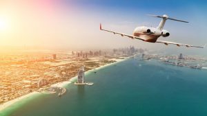 Un jet privé survolant Dubaï au coucher du soleil, avec une vue dégagée sur les gratte-ciel, l'océan et le bâtiment distinctif Burj Al Arab en forme de voile.