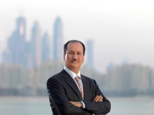 Un homme professionnel en costume se tient les bras croisés, avec un horizon flou de Dubaï en arrière-plan. Il a une expression neutre et porte un costume sombre avec une cravate rouge.