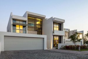Villa moderne à Dubaï avec un toit plat, des murs blancs et de grandes fenêtres éclairées au crépuscule. Un garage double et un jardin paysagé sont visibles devant.