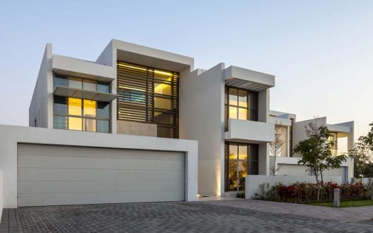 Villa moderne à Dubaï avec un toit plat, des murs blancs et de grandes fenêtres éclairées au crépuscule. Un garage double et un jardin paysagé sont visibles devant.