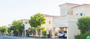 Une rue de banlieue calme bordée de maisons modernes à deux étages dotées de balcons et de portes cintrées, d'arbres verts luxuriants et de voitures garées sous un ciel clair se transforme en un quartier chic de Dubaï où les investisseurs