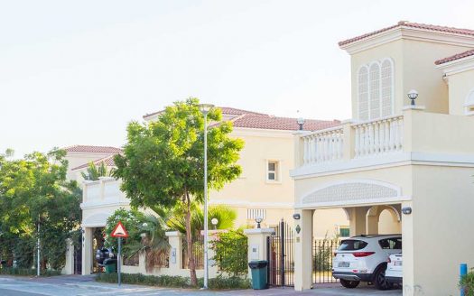 Une rue de banlieue calme bordée de maisons modernes à deux étages dotées de balcons et de portes cintrées, d'arbres verts luxuriants et de voitures garées sous un ciel clair se transforme en un quartier chic de Dubaï où les investisseurs