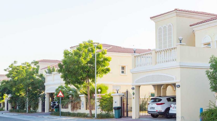 Une rue de banlieue calme bordée de maisons modernes à deux étages dotées de balcons et de portes cintrées, d&#039;arbres verts luxuriants et de voitures garées sous un ciel clair se transforme en un quartier chic de Dubaï où les investisseurs