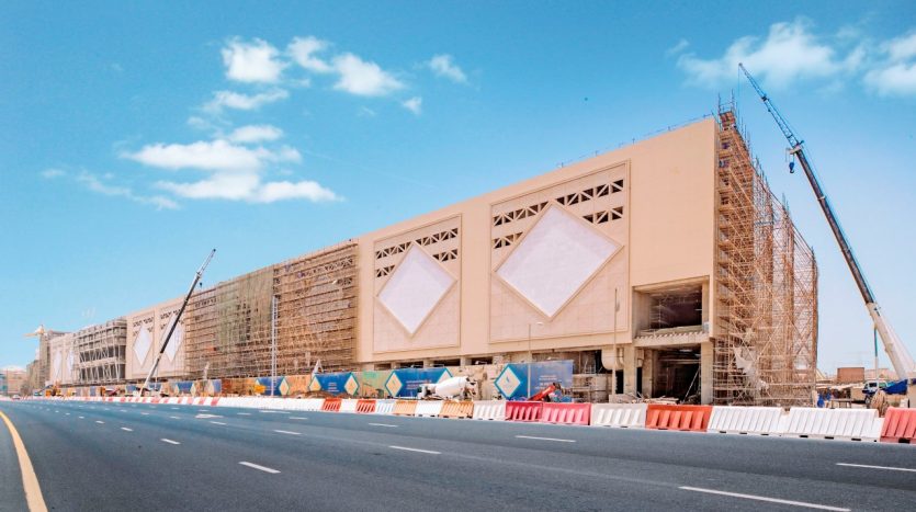 Un grand chantier de construction le long d&#039;une autoroute avec des grues et des échafaudages autour d&#039;un bâtiment beige partiellement achevé avec de grands motifs en forme de losange sur sa façade sous un ciel bleu clair à Dubaï.
