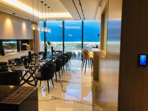 Cuisine de luxe moderne avec un coin repas doté de chaises sombres, d'une table élégante et de suspensions. Sol et armoires réfléchissants, avec de grandes fenêtres montrant le paysage extérieur dans un appartement haut de gamme à Dubaï.