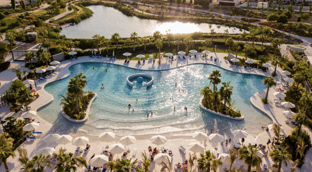 Vue aérienne d'une piscine animée de style complexe avec des gens nageant et se prélassant sous des parasols blancs, entourée d'une verdure luxuriante et d'un lac, parfaite pour ceux qui cherchent à investir à Dubaï