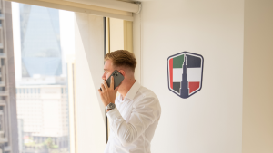 Un professionista in camicia bianca parla allo smartphone vicino alla finestra di un ufficio, con il logo di un'agenzia immobiliare di Dubai sulla parete accanto.