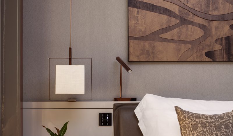 Un coin chambre confortable dans une propriété de l&#039;immobilier Dubaï comprenant un lit soigneusement fait avec des draps blancs et des oreillers décoratifs, une table de chevet avec une lampe et une œuvre d&#039;art abstraite en bois au mur.