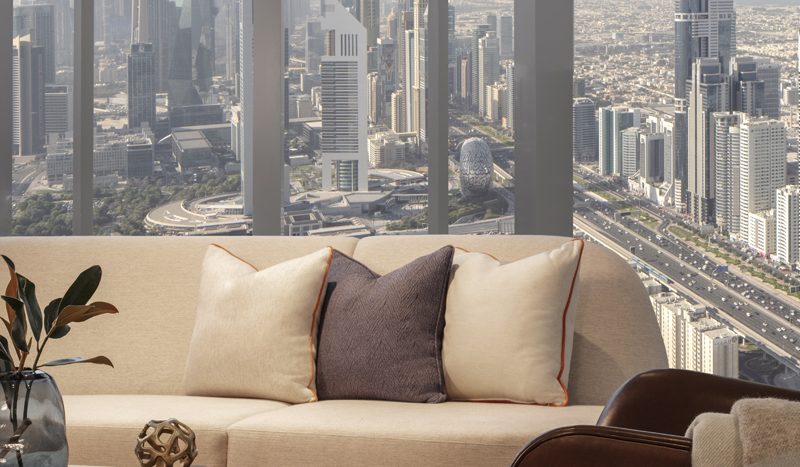 Un salon moderne dans une villa de Dubaï avec un canapé beige, des coussins et une couverture, doté de grandes fenêtres donnant sur une vue panoramique sur un paysage urbain avec des gratte-ciel sous un ciel bleu clair