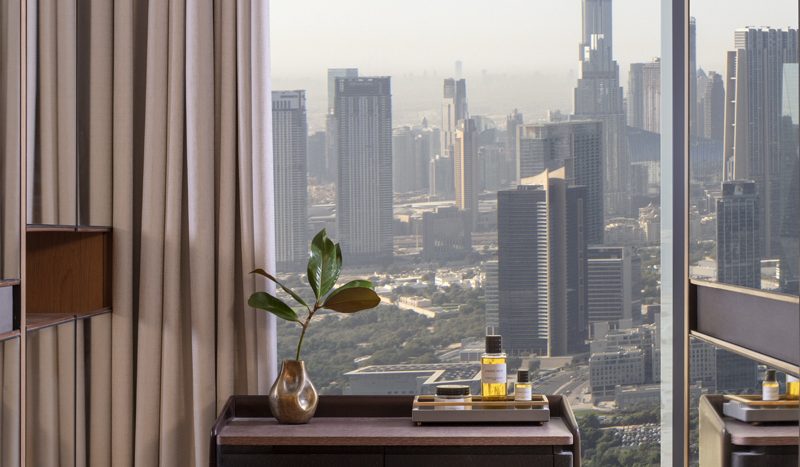 Un bureau moderne dans un appartement de Dubaï comprenant un bureau en bois et un petit tabouret, élégamment placés près d&#039;une grande fenêtre offrant une vue panoramique sur les toits de la ville avec des gratte-ciel, dont un imposant