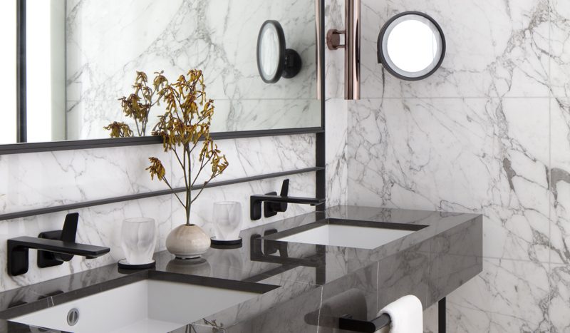 Une salle de bains luxueuse dans un appartement de Dubaï, avec des murs et un plan de vasque recouverts de marbre blanc, comprenant une douche aux parois de verre, un grand miroir et d&#039;élégants lavabos. Des serviettes aux tons neutres complètent le