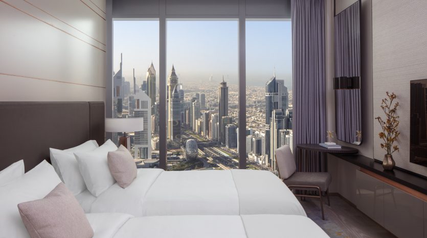 Chambre d&#039;hôtel luxueuse avec un grand lit face à une baie vitrée offrant une vue panoramique sur les toits de la ville moderne de Dubaï pendant la journée.