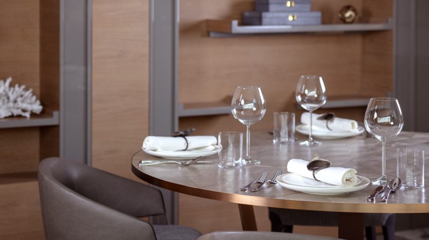Table à manger élégante avec assiettes blanches, serviettes pliées, verres à vin et couverts dans un appartement de Dubaï doté d&#039;un mobilier en bois et d&#039;un éclairage tamisé.
