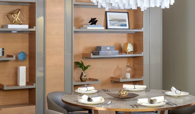 Une salle à manger moderne dans un appartement de Dubaï comprenant une table ronde en bois avec quatre chaises vert olive, complétées par un lustre blanc et cuivre, des armoires en bois et des objets décoratifs.