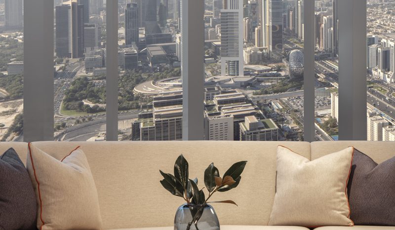 Un salon moderne avec un grand canapé et une table basse ornée d&#039;une plante dans un vase, offrant une vue panoramique sur les toits de Dubaï à travers des baies vitrées.