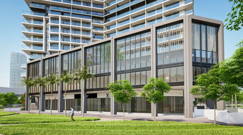 Un bâtiment moderne à plusieurs étages avec de grandes fenêtres vitrées aux étages inférieurs et des espaces de vente au détail. Une herbe verte luxuriante entoure la structure, sous un ciel clair à Dubaï.