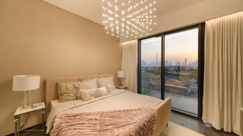 Chambre d&#039;appartement moderne à Dubaï avec un grand lit recouvert d&#039;une literie rose tendre, un lustre élégant et une baie vitrée montrant les toits de la ville au coucher du soleil.
