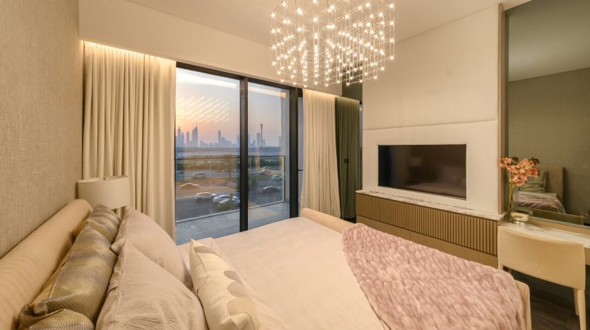 Chambre moderne avec de grandes fenêtres mettant en valeur les toits de la ville, un lit moelleux avec une couverture rose, une télévision montée et un lustre saisissant, créant une ambiance chaleureuse dans une propriété d&#039;investissement à Dubaï.