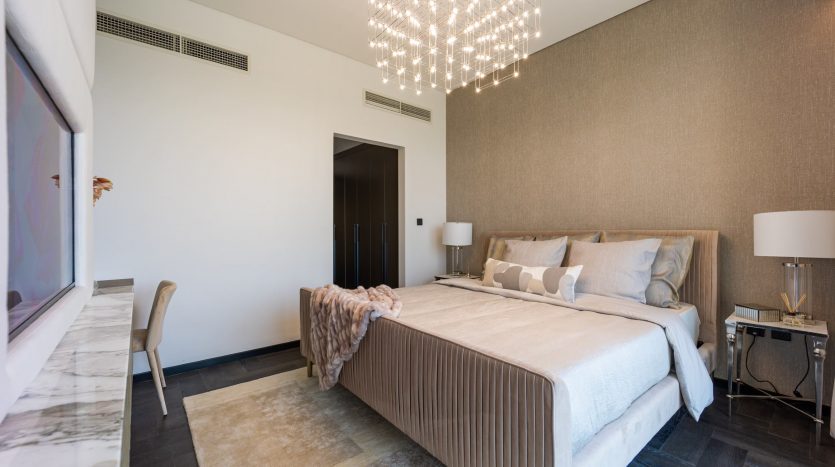 Une chambre moderne dans une villa de Dubaï comprenant un grand lit avec des draps beiges, un lustre élégant, des murs texturés, un tapis moelleux et un mobilier contemporain, baigné de lumière naturelle.