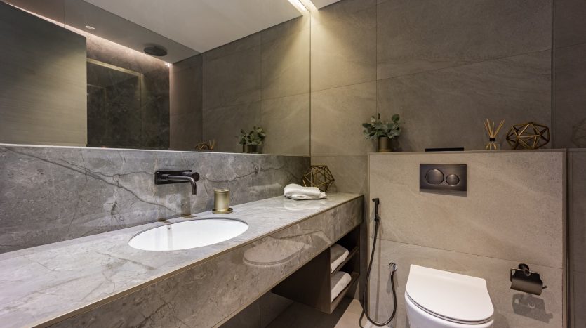 Salle de bains moderne dans un appartement de Dubaï avec murs et comptoirs en marbre gris, lavabo intégré, grand miroir et toilettes. Le décor comprend de petites plantes vertes et des accents géométriques. Design minimaliste avec éclairage ambiant.