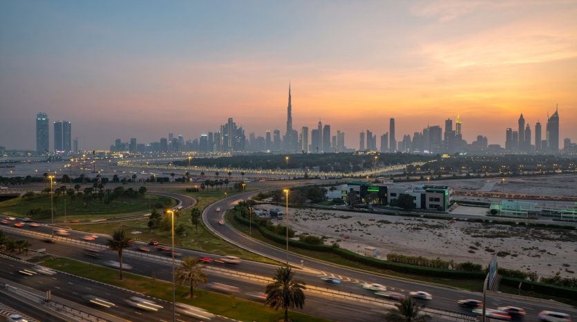 Vue au coucher du soleil sur une ville animée avec des gratte-ciel visibles, dont une haute tour à Dubaï, avec des autoroutes éclairées et un ciel orange vif en arrière-plan.