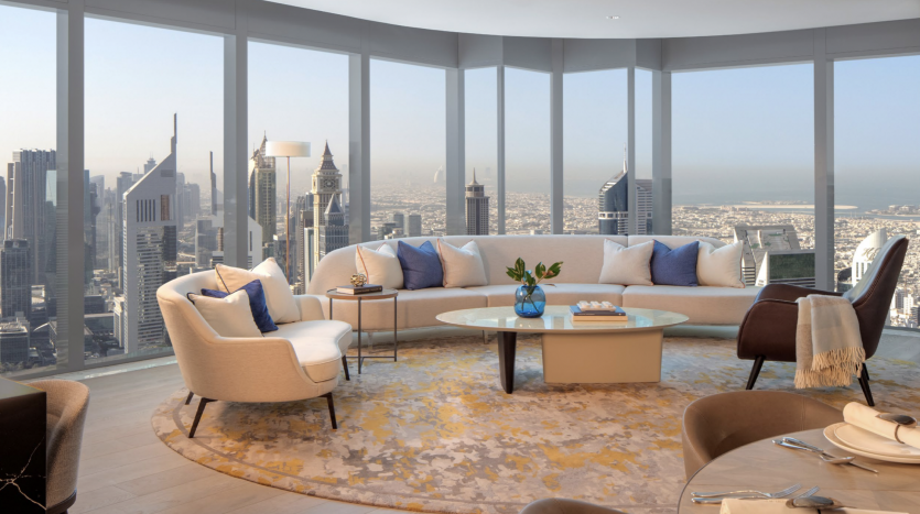 Salon luxueux dans un appartement de Dubaï avec un mobilier moderne et de grandes fenêtres panoramiques offrant une vue imprenable sur les toits de la ville. Le décor comprend une table basse circulaire, des canapés et des chaises.