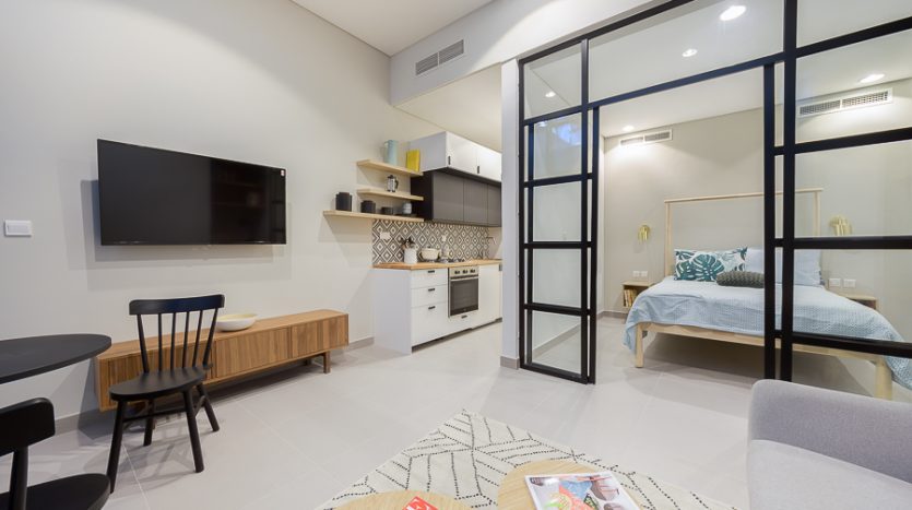 Intérieur d'un studio moderne à Dubaï avec un salon et une chambre connectés, avec des cloisons en verre à cadre noir, un mobilier minimaliste, une télévision murale et une kitchenette.