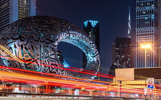Paysage urbain nocturne montrant le musée du futur à Dubaï avec une calligraphie arabe complexe sur sa structure, des traînées de lumière vibrantes provenant de véhicules et des gratte-ciel illuminés en arrière-plan mettant en vedette un appartement de premier plan à Dubaï