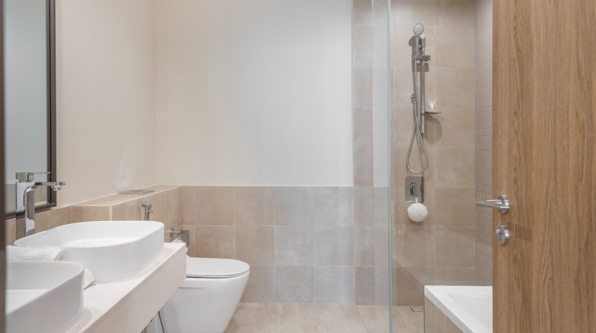 Une salle de bains moderne comprenant un lavabo blanc, des toilettes et une douche à l&#039;italienne avec portes vitrées, le tout sur des murs et un sol carrelés beiges. Les armoires en bois naturel ajoutent de la chaleur à la villa à Dubaï.