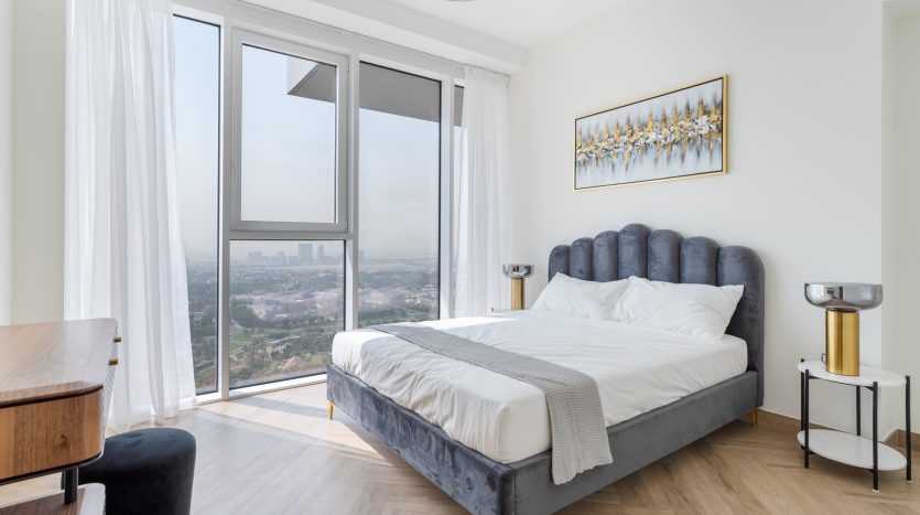Une chambre moderne et ensoleillée comprenant un lit rembourré gris moelleux avec une literie blanche, une baie vitrée avec des rideaux transparents, un mobilier minimaliste et une vue panoramique sur la ville depuis une villa.