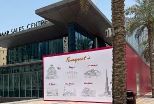 Un panneau publicitaire pour Fauchon Paris présentant des croquis de lieux emblématiques, dont Paris et New York, devant un immeuble moderne avec un palmier sur le côté, sous un ciel bleu éclatant à Dubaï