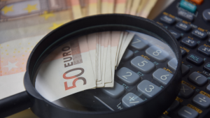 Une loupe posée sur une pile de billets de 50 euros, destinés à Investissement Dubaï, partiellement visibles en dessous, avec une calculatrice floue en arrière-plan sur un bureau.