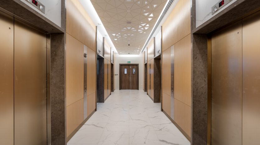 Un hall d&#039;ascenseur moderne dans une villa de Dubaï comprenant quatre ascenseurs aux finitions en bois et métalliques, des sols en marbre blanc et un plafond à motifs géométriques. La lumière est répartie uniformément le long du couloir.