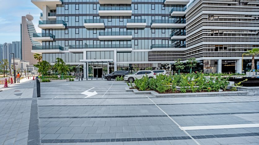 Un paysage urbain moderne à Dubaï avec un trottoir à motifs géométriques, des jardinières vertes luxuriantes et des bâtiments élégants avec des balcons horizontaux. Une voiture de luxe est visible près de l&#039;entrée de l&#039;un
