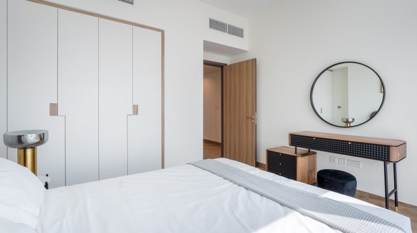 Une chambre moderne et minimaliste dans une villa de Dubaï avec un grand lit centré dans le cadre, flanqué d&#039;une console en bois et métal noir et d&#039;un miroir rond au mur. Lumineux, neutre