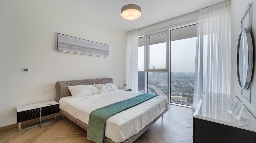 Une chambre moderne dans un appartement à Dubaï avec un grand lit, une literie blanche et un jeté vert. Grande fenêtre avec vue sur la ville, décoration minimaliste et parquet.
