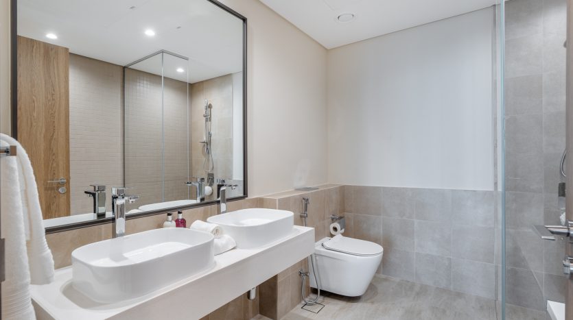 Intérieur de salle de bains moderne dans un appartement de Dubaï comprenant deux lavabos, un grand miroir et des toilettes. Tons neutres avec murs carrelés et détails d&#039;armoires en bois.